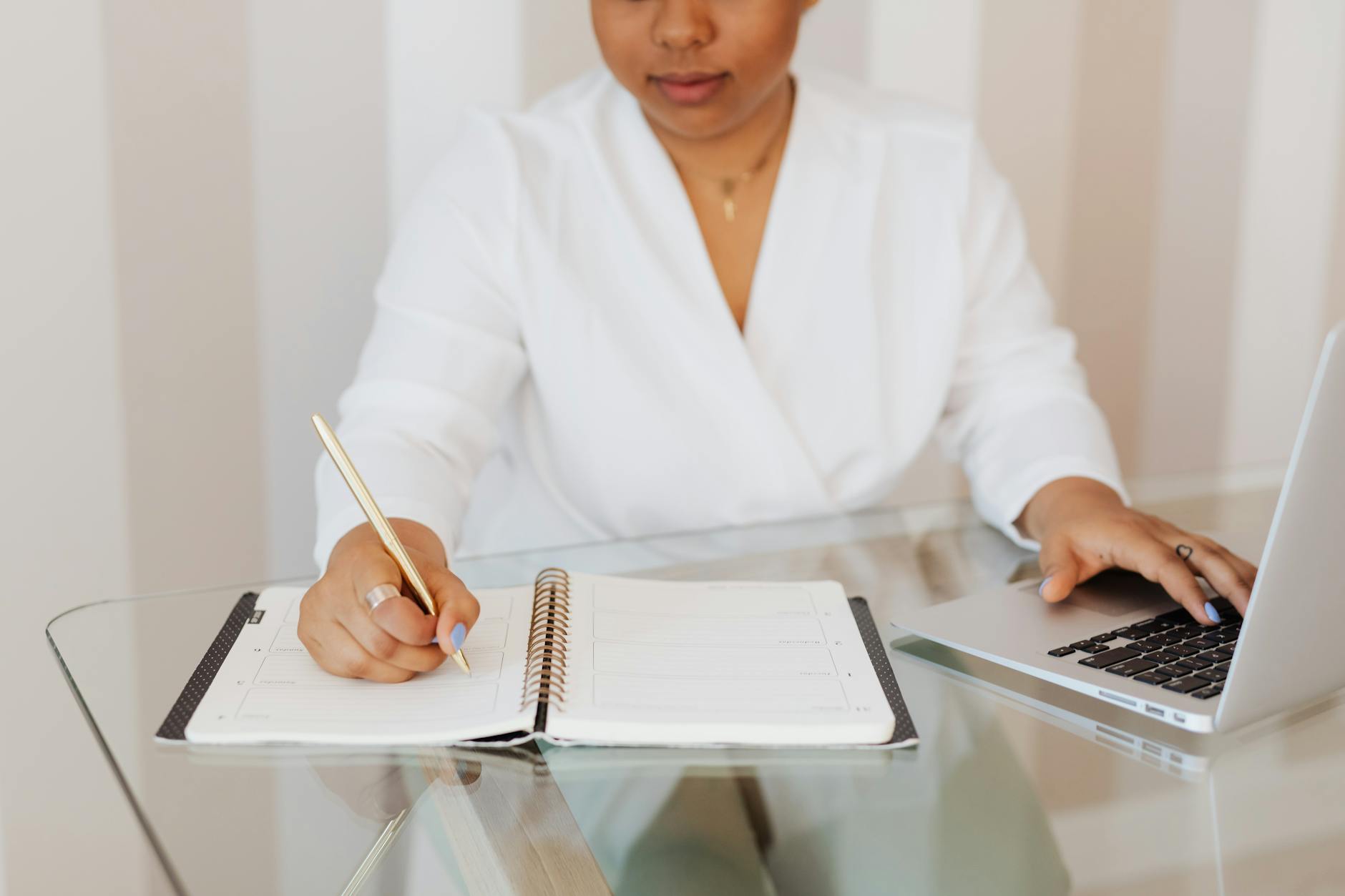 person in white top writing on notebook while using a laptop